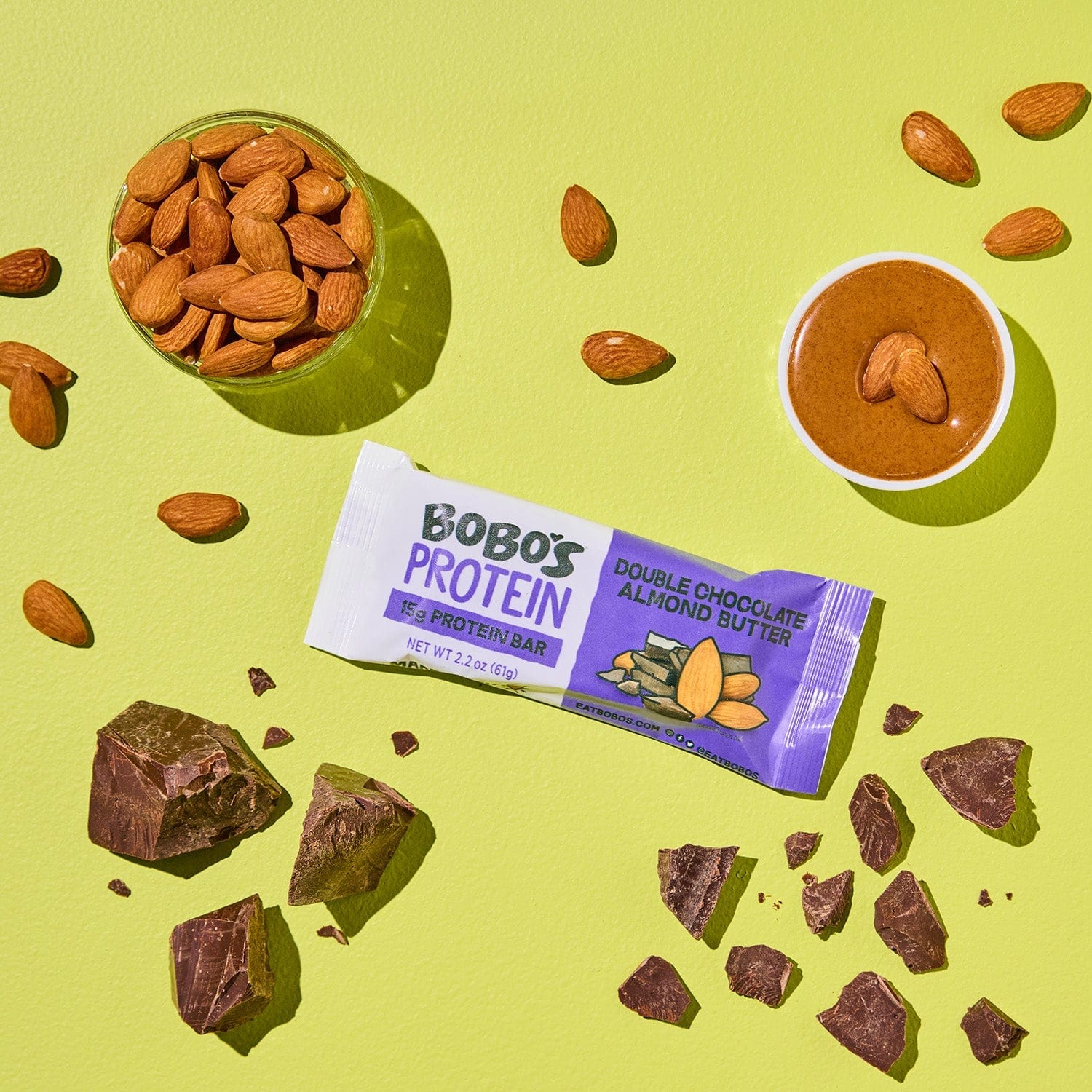 Protein bar on green background with surrounding chocolate chunks and almond butter