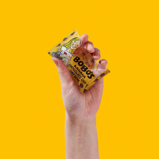 A hand holds a packaged snack labeled "Bobo's Banana Chocolate Chip Oat Bite" against a bright yellow background.