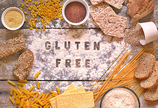 Various pastas and grains surrounding a floured wooden surface with "Gluten Free" written in the flour