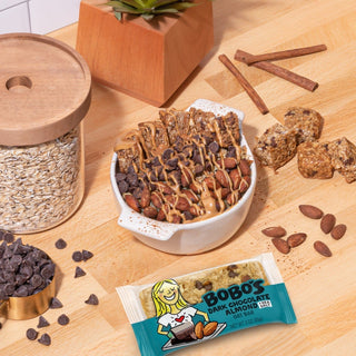 A bowl of Dark Chocolate Almond with Sea Salt Oat Bar, featuring oats, chocolate chips, and cinnamon sticks on a table with a jar of oats nearby.