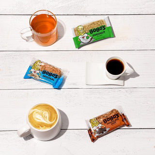 A group of coffee cups, a glass cup of liquid, a swirl of foam, and oat bars on a white table. Bobo’s Original Oat Bar